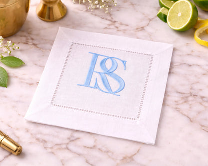 White cocktail napkin with blue monogram 'RS' on a marble surface with limes and flowers.