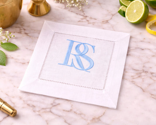 White cocktail napkin with blue monogram 'RS' on a marble surface with limes and flowers.