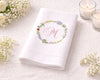 White folded napkin with floral monogram design on a white surface with candles and flowers.