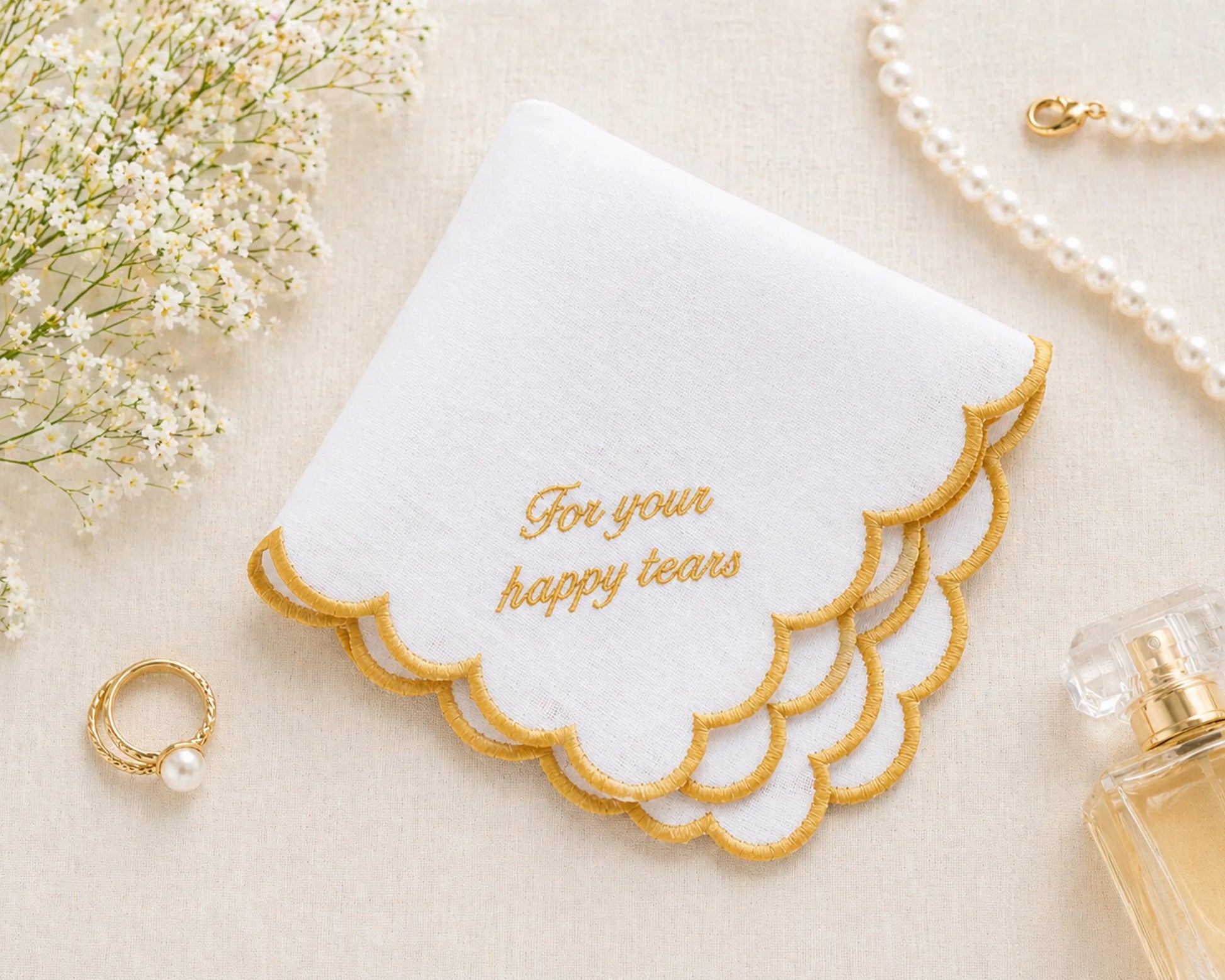 White handkerchief for bride with gold text 'For your happy tears' on a beige surface with flowers and a ring.