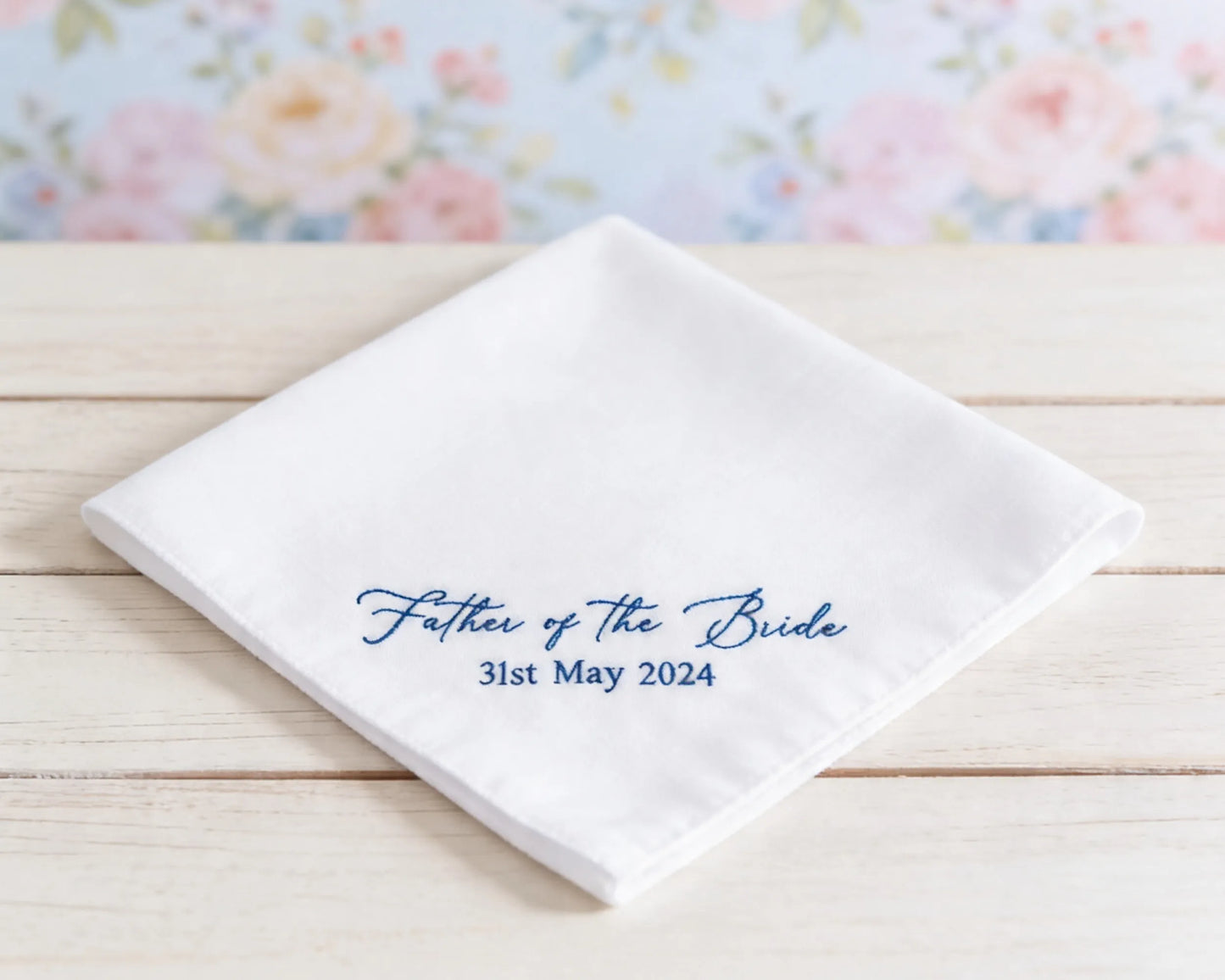 White handkerchief with 'Father of the Bride' text on a wooden surface with floral background