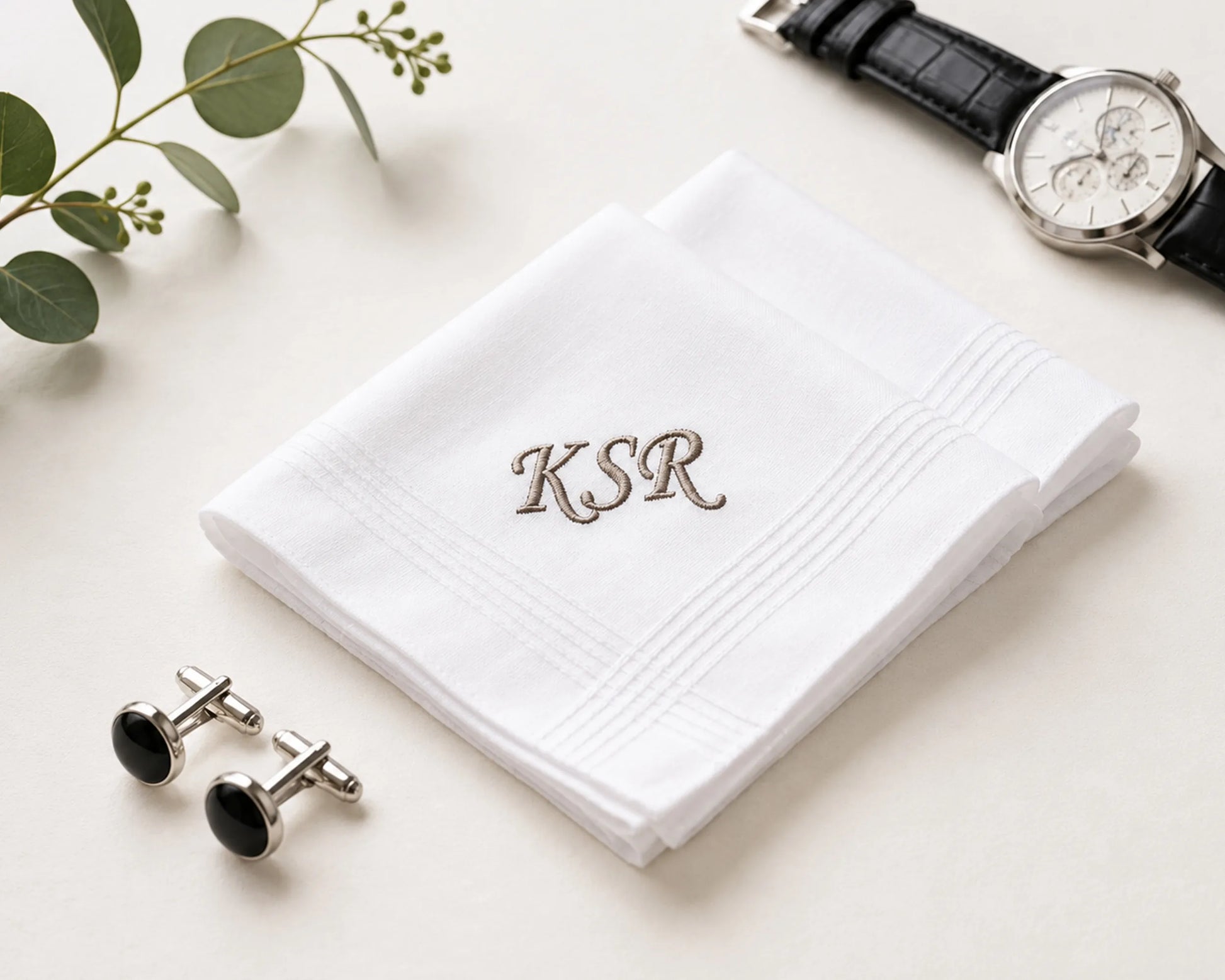 White handkerchief with 'KSR' embroidery, cufflinks, and a watch on a light surface.
