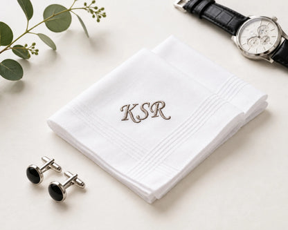 White handkerchief with 'KSR' embroidery, cufflinks, and a watch on a light surface.