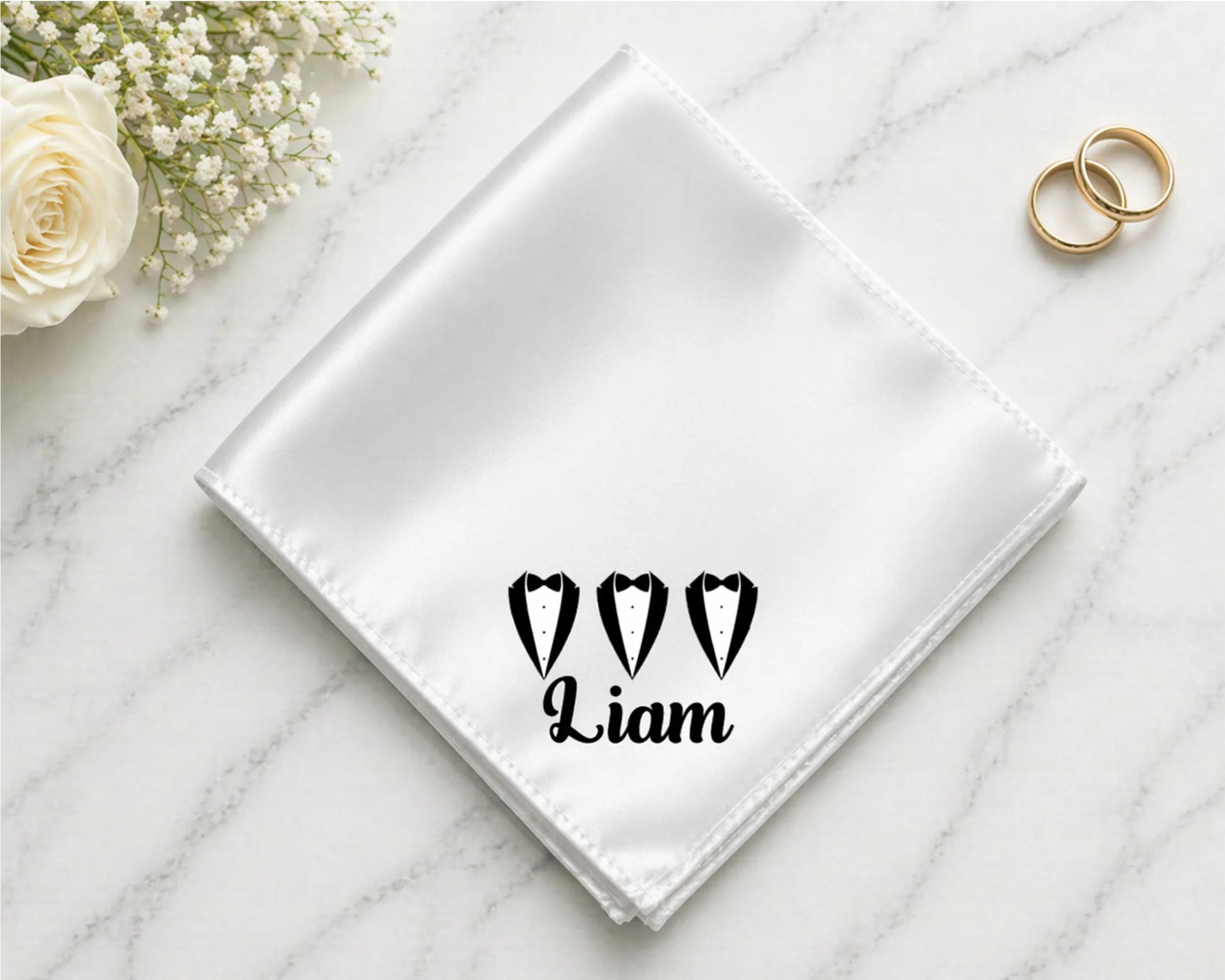 White handkerchief with 'Liam' and heart designs on a marble surface with flowers and rings.