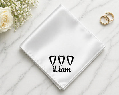 White handkerchief with 'Liam' and heart designs on a marble surface with flowers and rings.