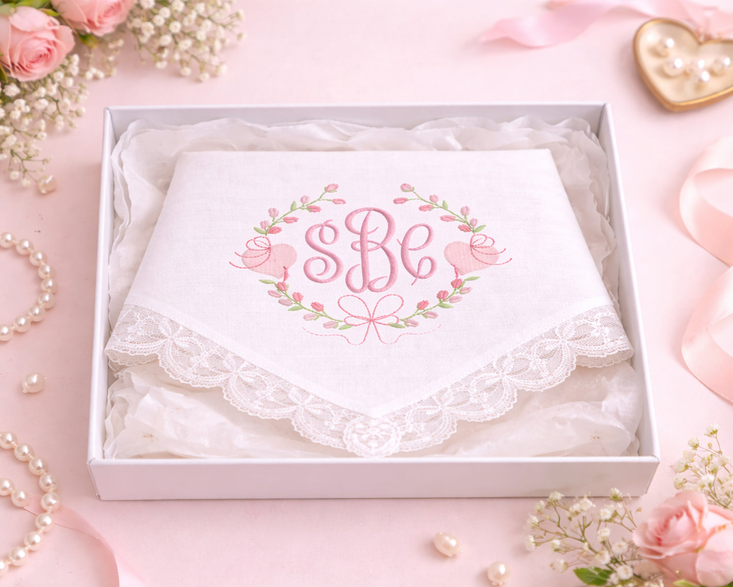 White handkerchief with 'SBe' monogram in a decorative box on a pink background with flowers and jewelry.