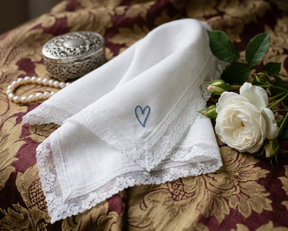 White handkerchief with a blue heart embroidery on a patterned fabric background with pearls and a flower.