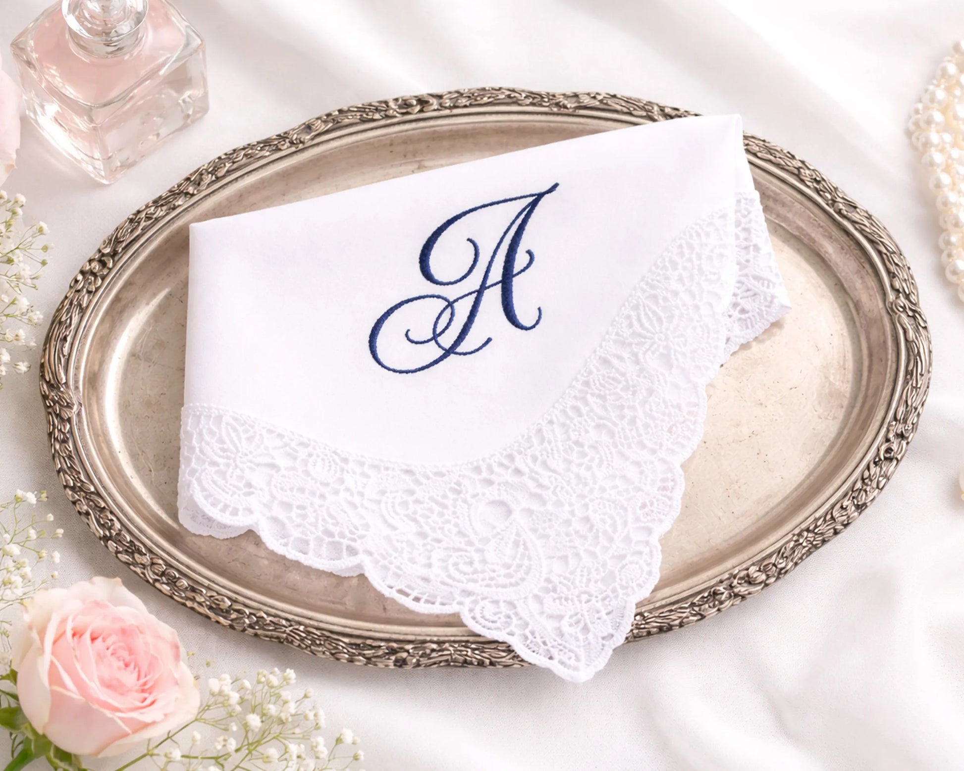 White handkerchief with a blue monogram 'A' on a decorative silver tray with flowers in the background.