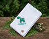 White handkerchief with a green dinosaur design and 'James' text, placed on a wooden surface with ferns and stones.