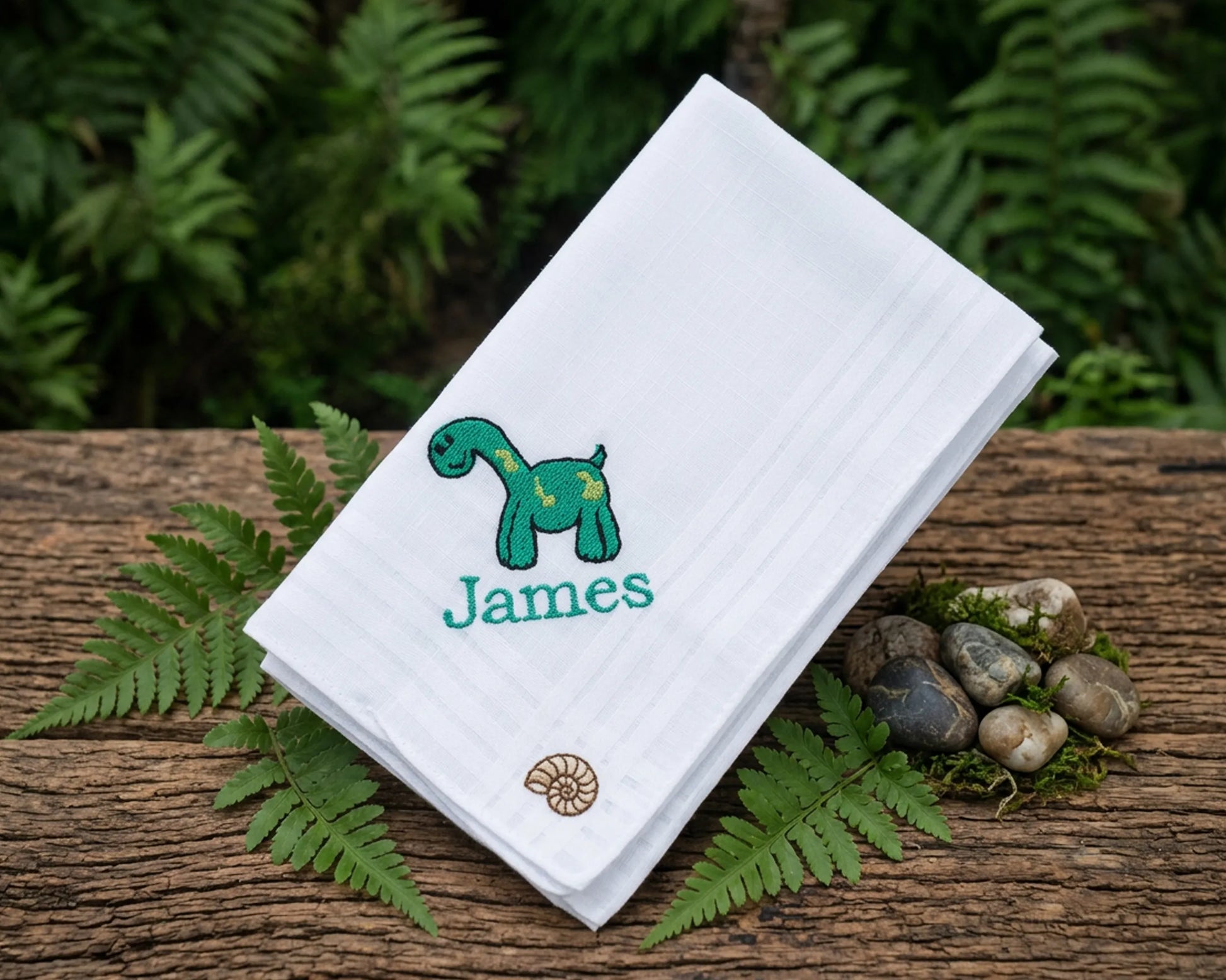 White handkerchief with a green dinosaur design and 'James' text, placed on a wooden surface with ferns and stones.