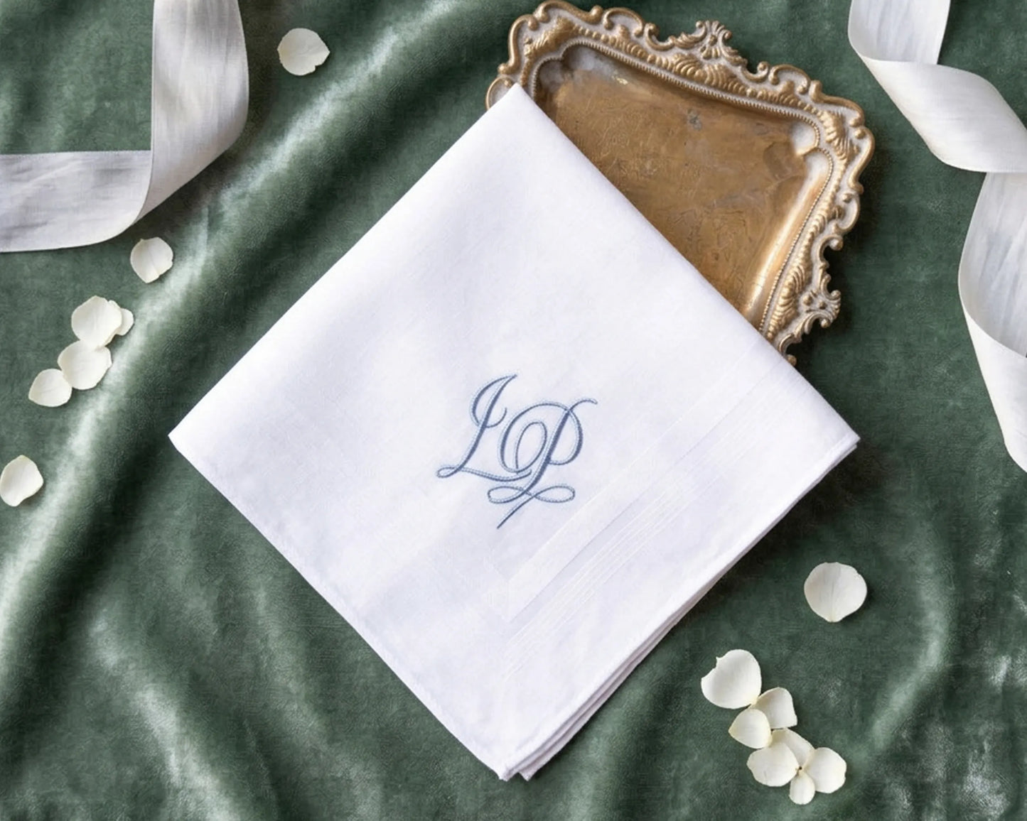 White handkerchief with blue monogram on a green fabric background with ribbons and petals.