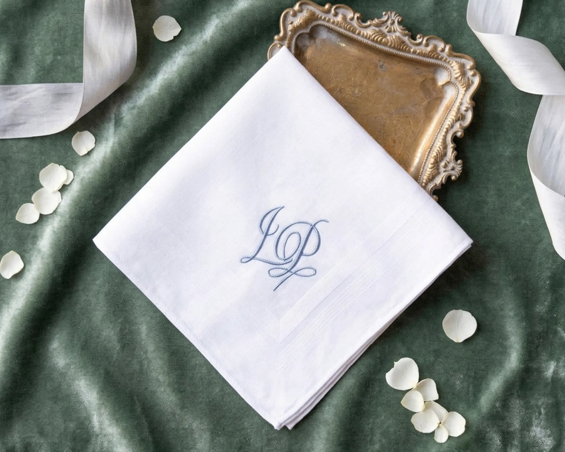 White handkerchief with blue monogram on a green fabric background with ribbons and petals.