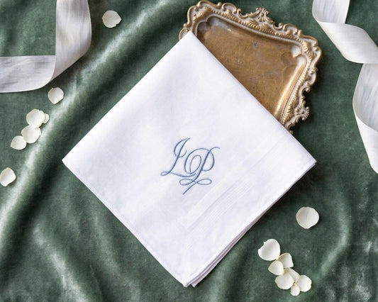 White handkerchief with blue monogram on a green fabric background with ribbons and petals.