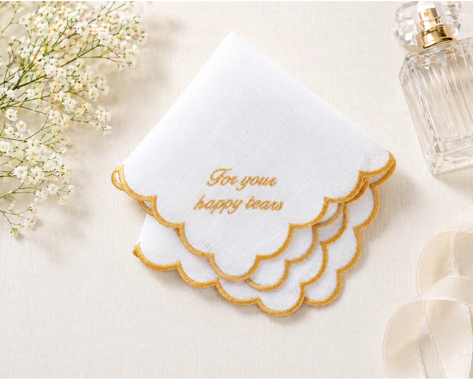 White handkerchief with gold text 'For your happy tears' on a light background with flowers and a perfume bottle.