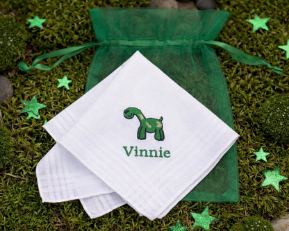 White handkerchief with green dinosaur design and 'Vinnie' text, placed on green fabric with stars on a grassy background.