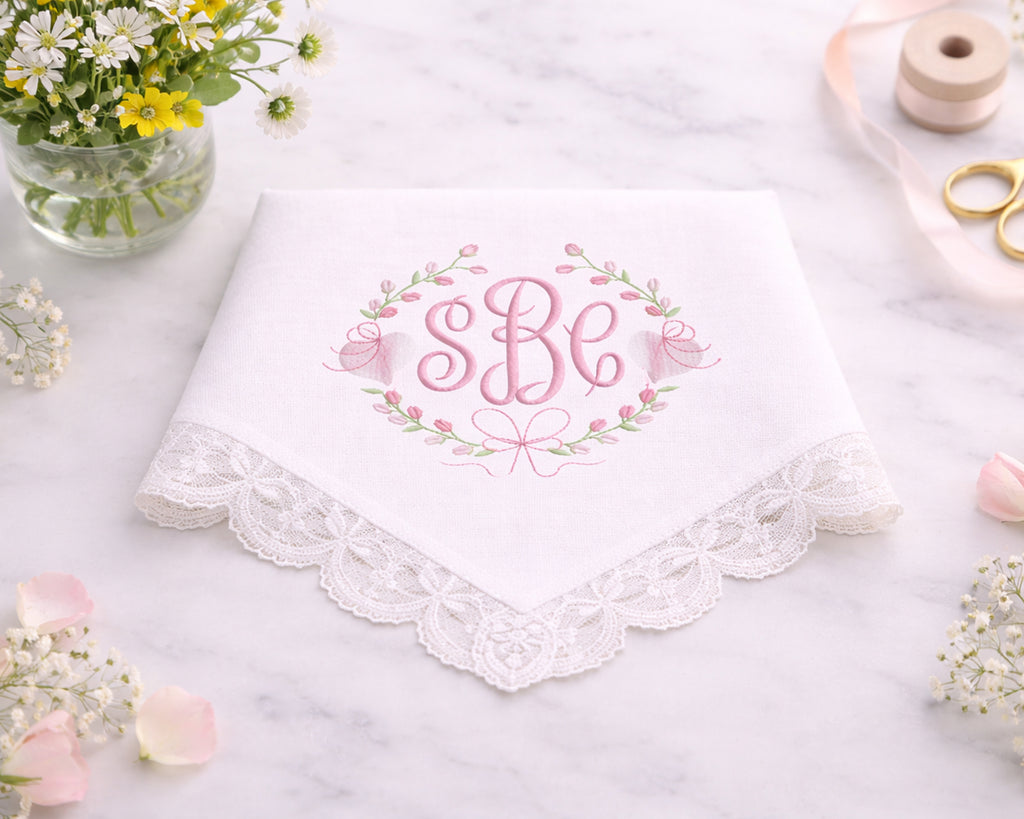 White handkerchief with lace trim and embroidered initials on a marble surface with flowers and sewing tools.
