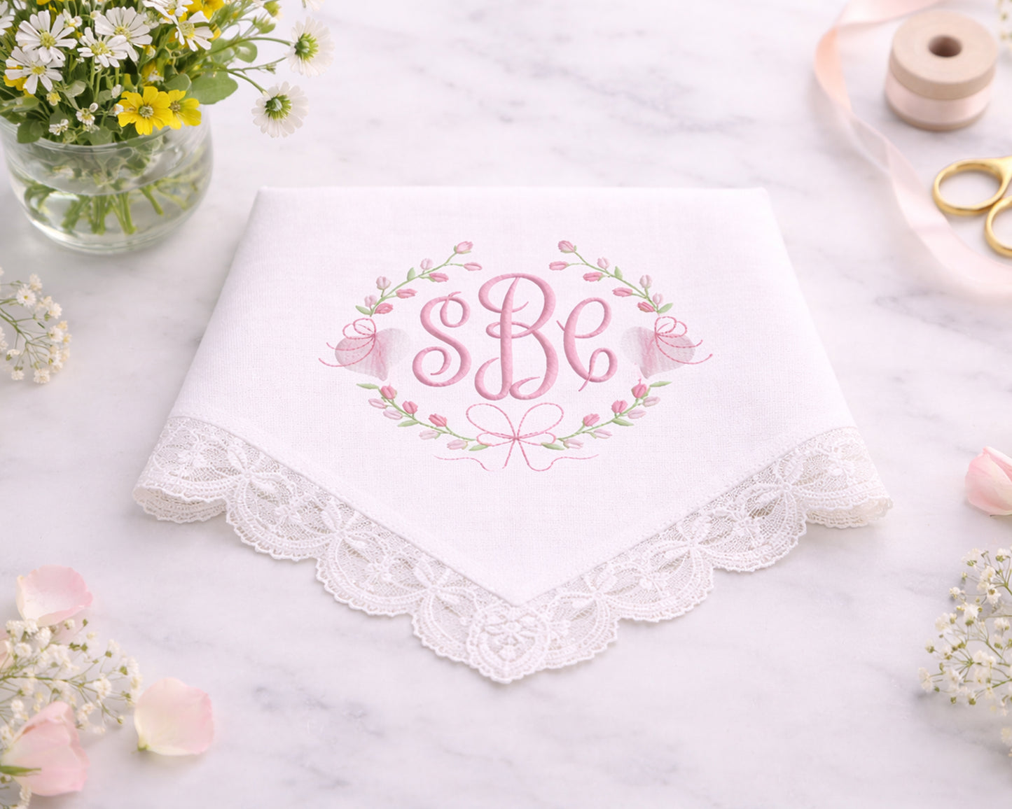 White handkerchief with lace trim and embroidered initials on a marble surface with flowers and sewing tools.