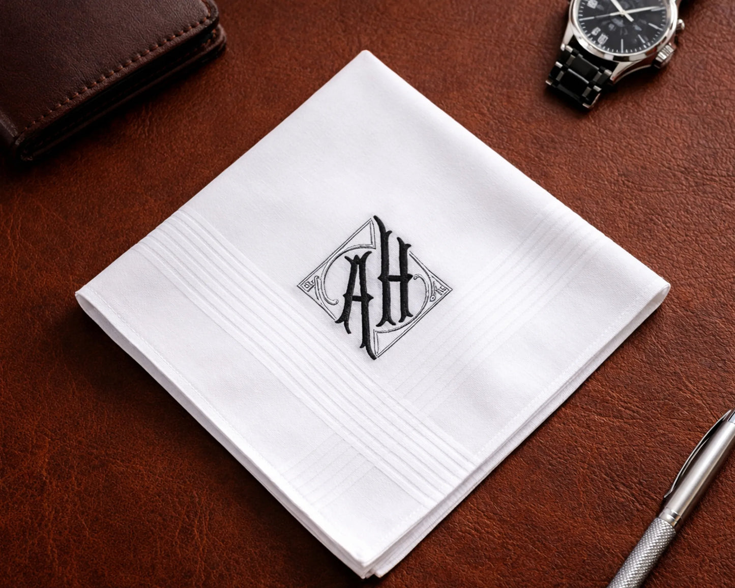 White handkerchief with monogram 'AH' on a brown leather surface with a pen and wallet.