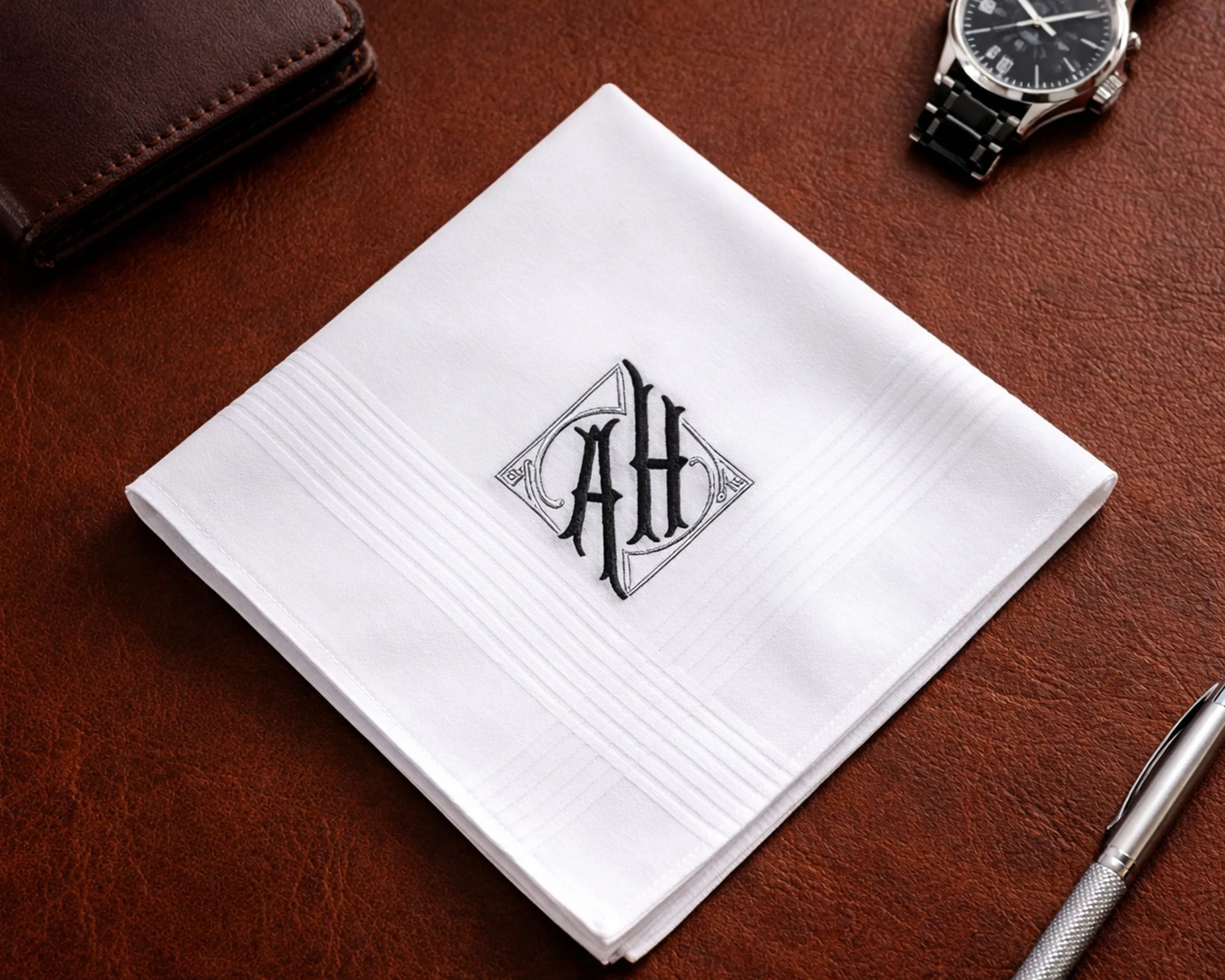White handkerchief with monogram 'AH' on a brown leather surface with a pen and wallet.