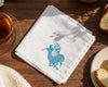 White hankie with a blue embroidered design on a wooden table with tea and bread.