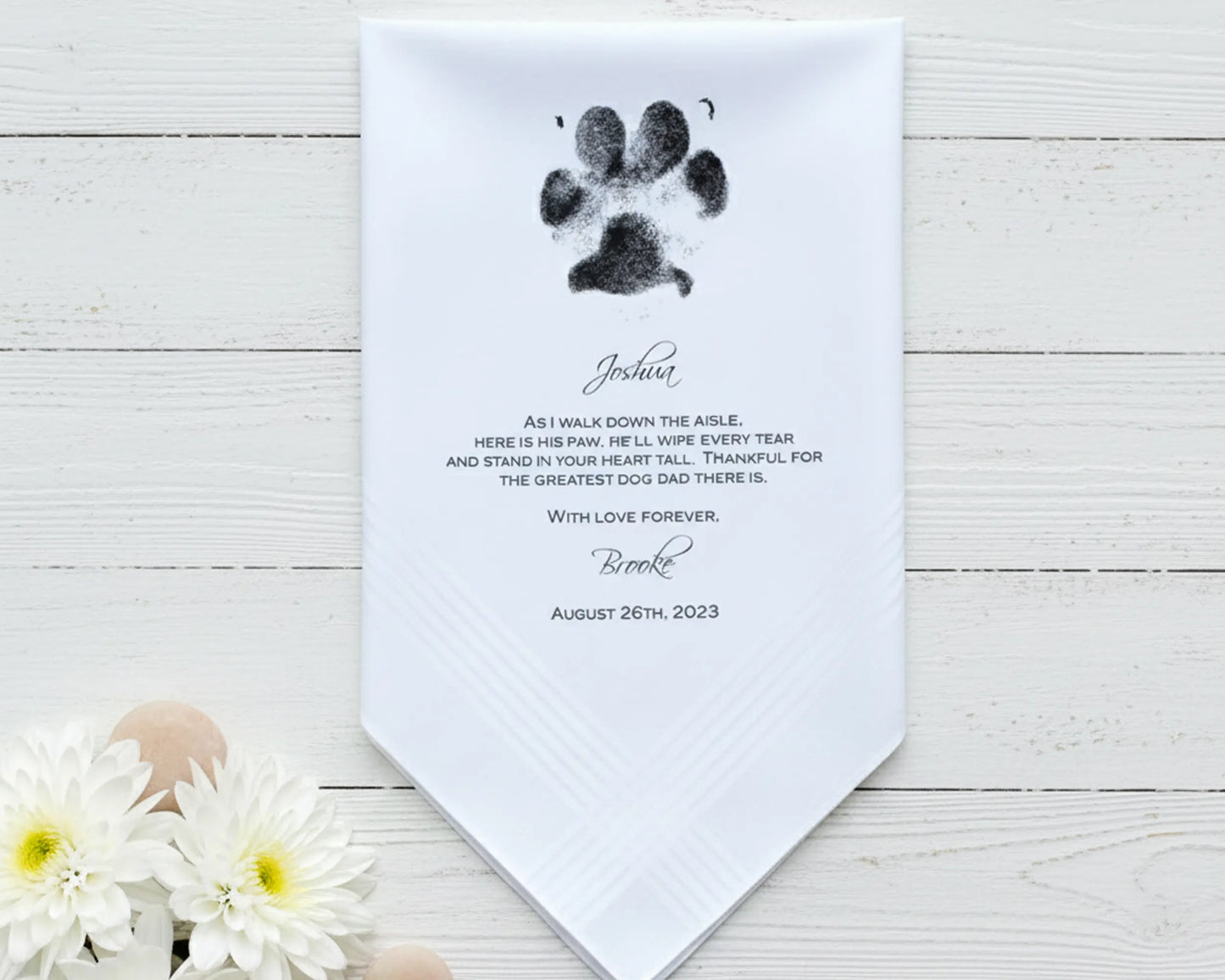 White hankiy with dog paw print and personalized text on a wooden surface with flowers.