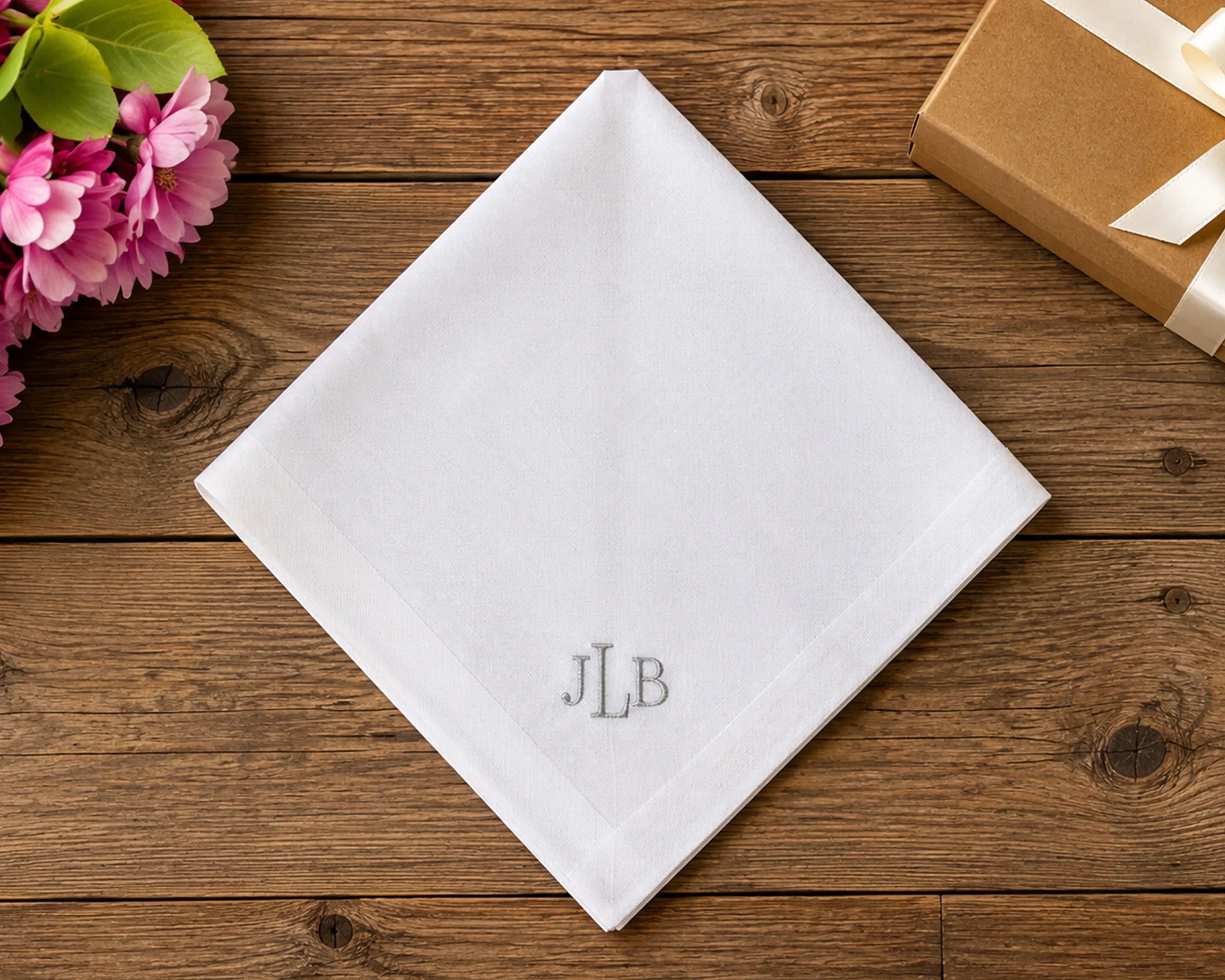 White hanky with 'JLB' monogram on a wooden surface with flowers and a gift box.