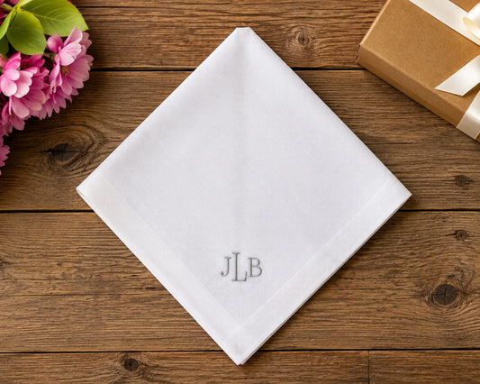 White hanky with 'JLB' monogram on a wooden surface with flowers and a gift box.