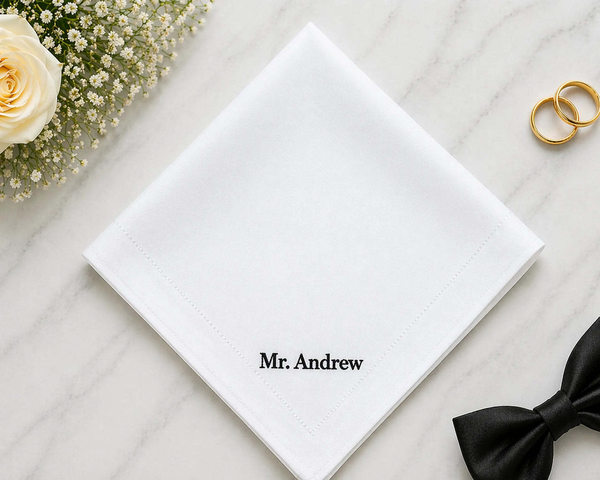 White hanky with 'Mr. Andrew' on it, accompanied by a black bow tie and gold rings on a light wooden surface.