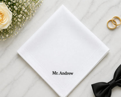 White hanky with 'Mr. Andrew' on it, accompanied by a black bow tie and gold rings on a light wooden surface.