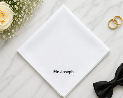White hanky with 'Mr. Joseph' text, gold rings, and a black bow tie on a marble surface with flowers.