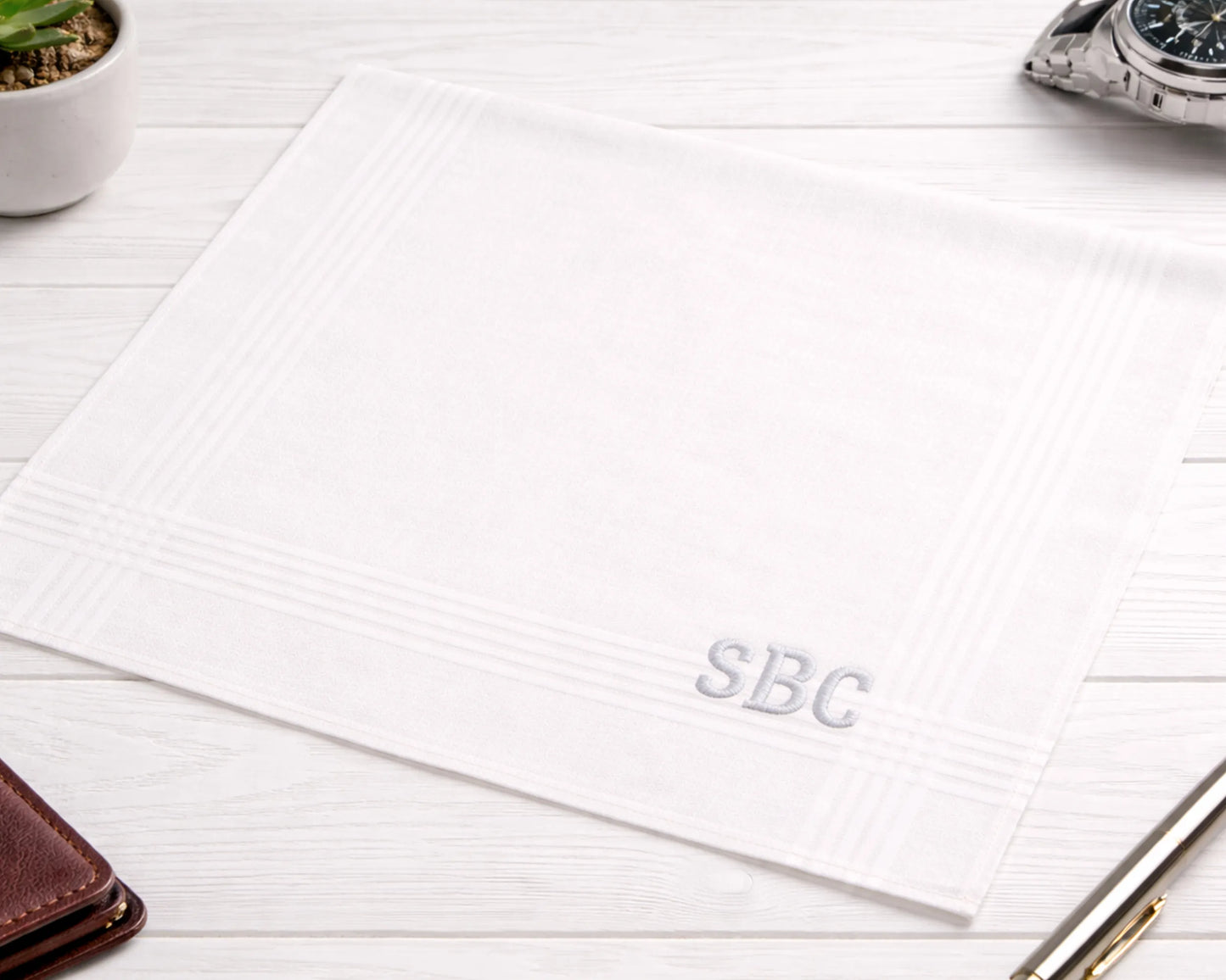 White hanky with 'SBC' initials on a wooden surface with a pen and watch.