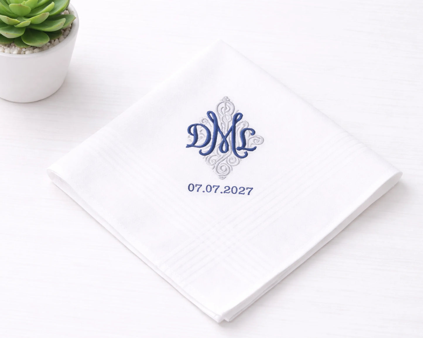 White hanky with blue monogram and date on a white surface with a small plant.