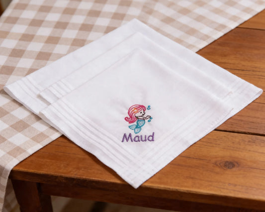 White hanky with embroidered mermaid design and 'Maud' text on a wooden table.