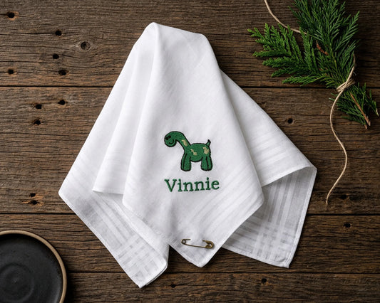 White hanky with green embroidered dinosaur and 'Vinnie' text on a wooden surface with greenery.
