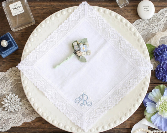 White lace handkerchief with embroidered letter 'R' on a wooden surface with flowers and a Chanel bottle.