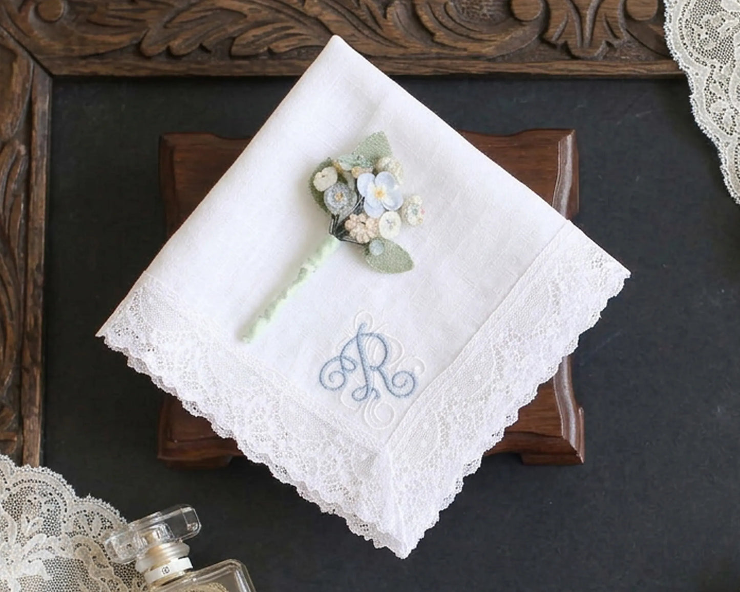 White lace handkerchief with floral arrangement and 'R' monogram on a dark surface.