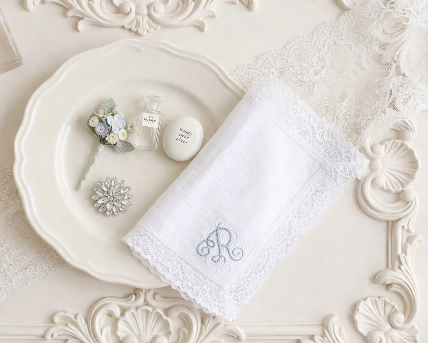 White lace handkerchief with monogram on a decorative white plate with small flowers and a bottle.