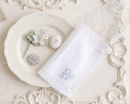 White lace handkerchief with monogram on a decorative white plate with small flowers and a bottle.
