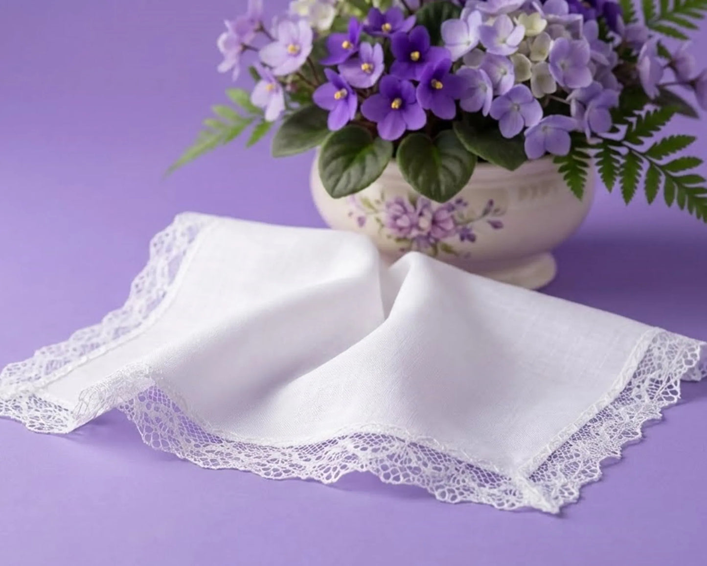 White lace-trimmed handkerchiefs on a purple surface with a vase of purple flowers.
