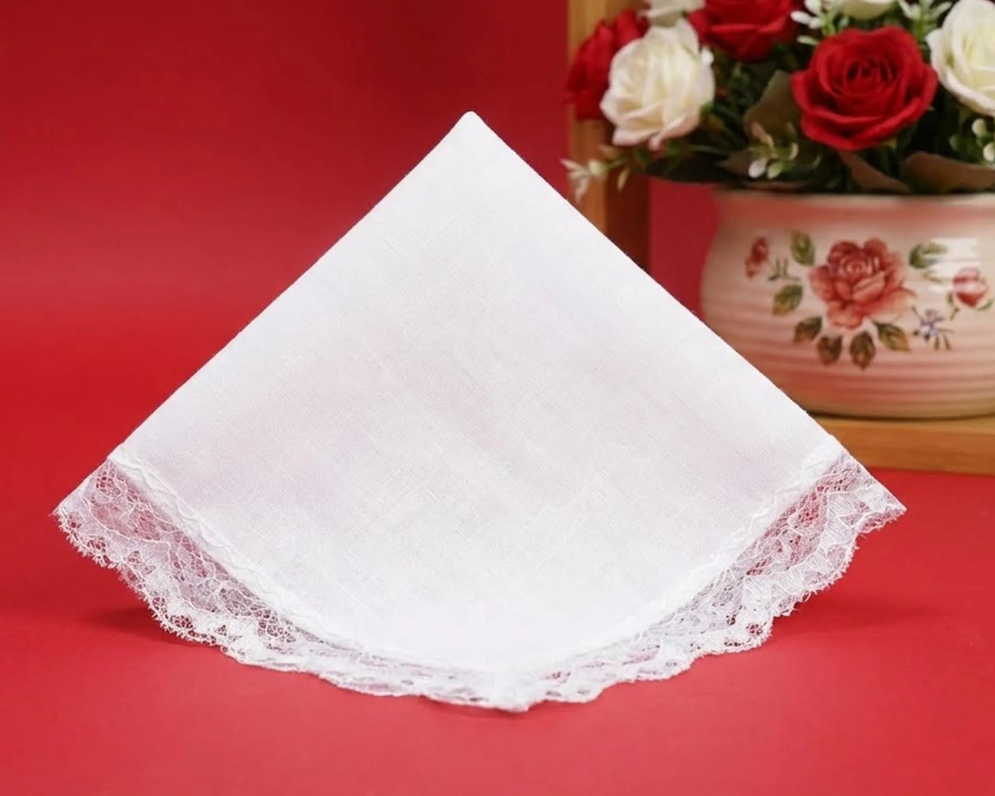 White lace-trimmed handkerchief on a red background with a floral arrangement in the background.