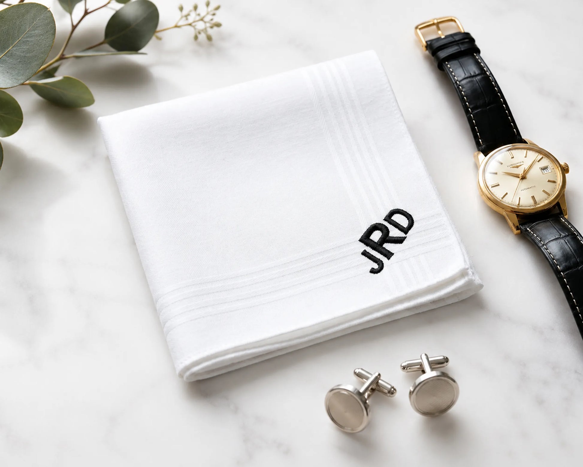 White handkerchief with initials, watch, and cufflinks on a marble surface.