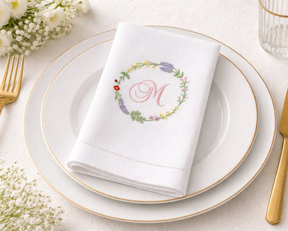 White napkin with floral design and monogram on a white plate with gold rim, surrounded by gold cutlery and flowers.
