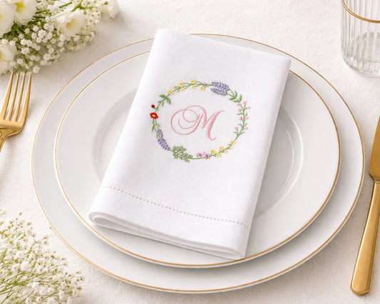 White napkin with floral design and monogram on a white plate with gold rim, surrounded by gold cutlery and flowers.