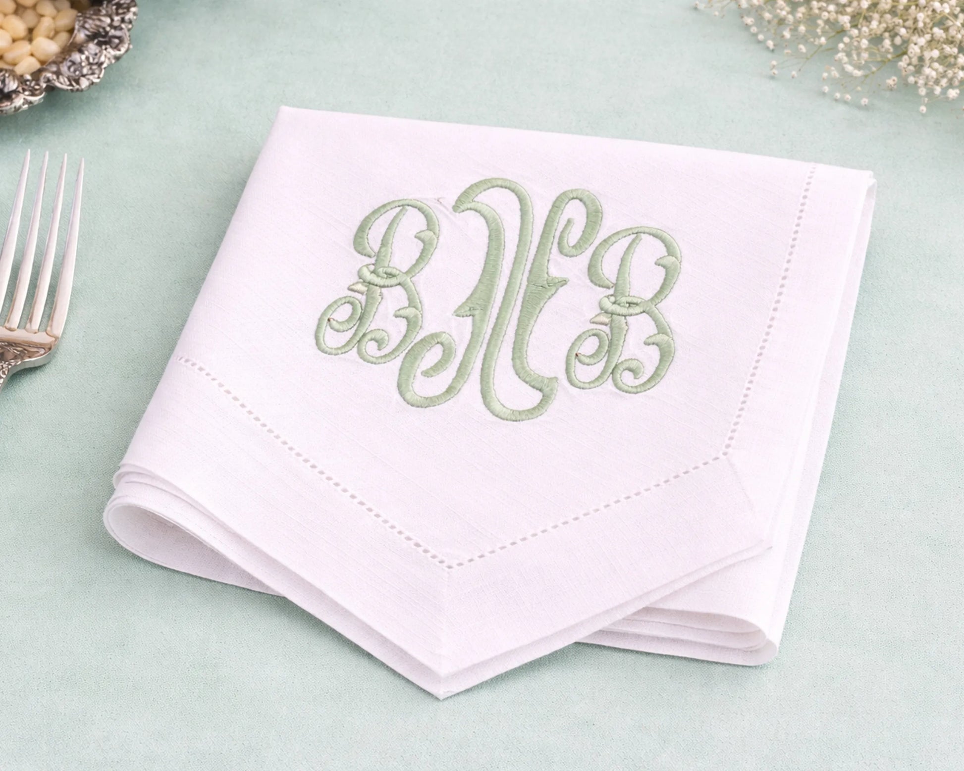 White napkin with green monogram on a light green surface with a fork and decorative elements.