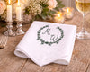 White napkin with monogram and wreath design on a wooden table with candles and a glass of wine.