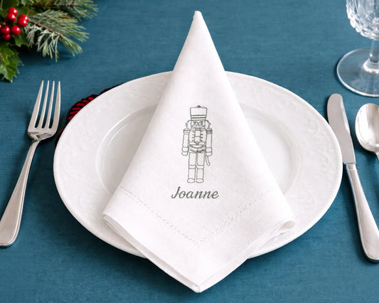 White plate with folded napkin featuring a nutcracker design and 'Joanne' text, on a blue tablecloth with Christmas decor.