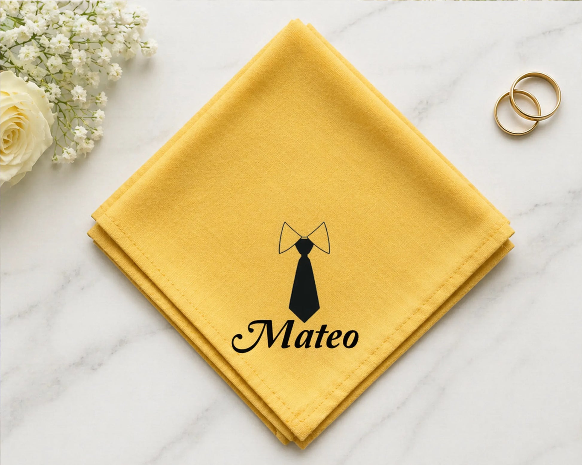 Yellow hanky with a black tie design and 'Mateo' text on a marble surface with flowers and rings.