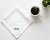 Flat lay of a white cotton handkerchief featuring bold black HJD initials next to a cup of black coffee.