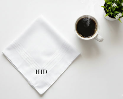 Flat lay of a white cotton handkerchief featuring bold black HJD initials next to a cup of black coffee.