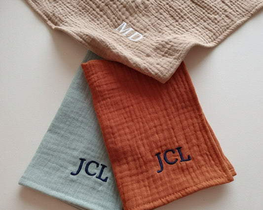 Two folded muslin cotton handkerchiefs in sage green and rust orange with navy JCL monogram embroidery.